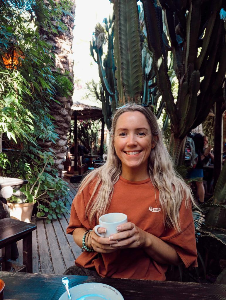 Jane in a rusty orange t shirt, smiling holding a coffee amongst the cactuses in La Sombra, Fuerteventura.