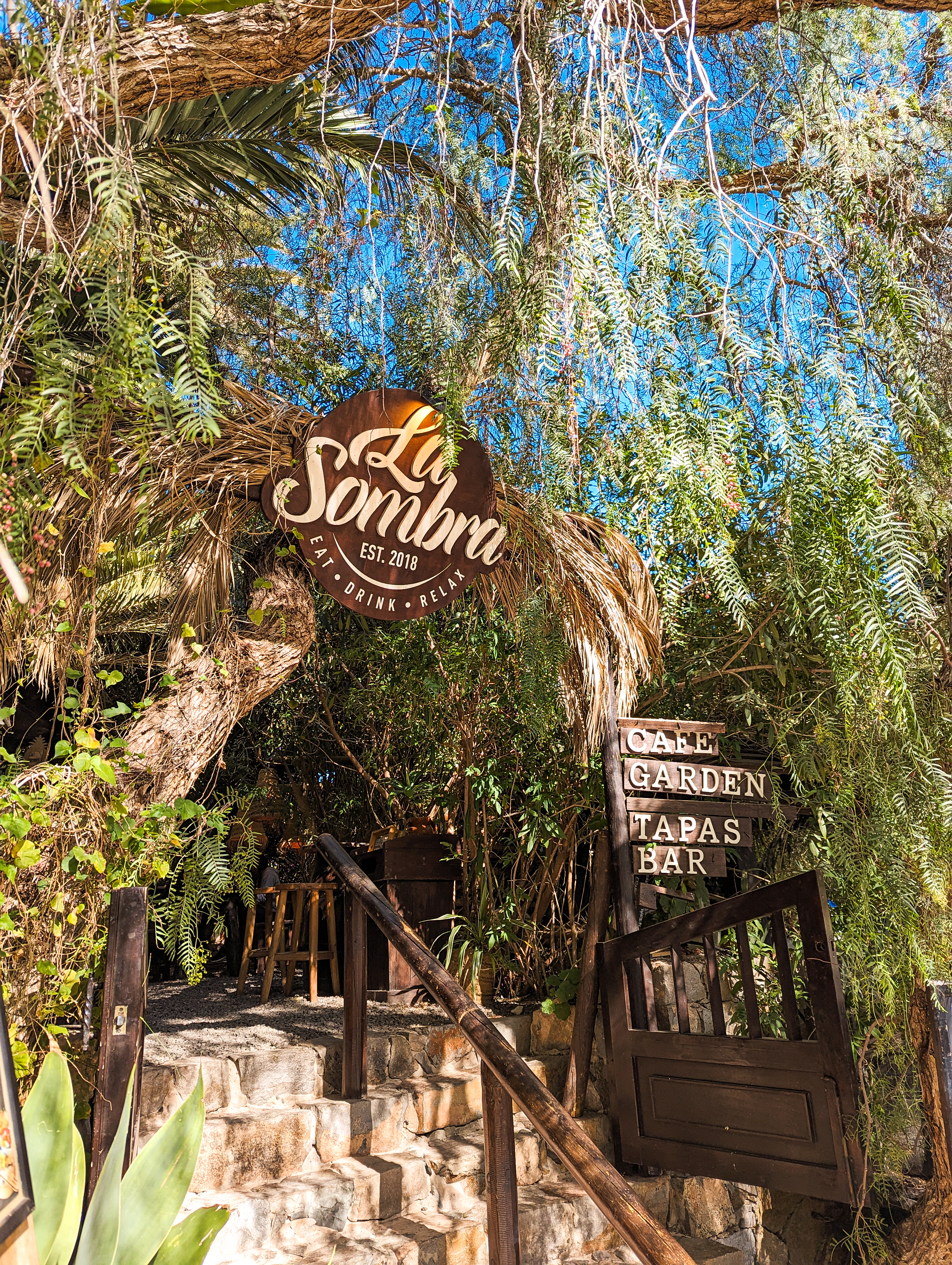 A picture of the entrance to La Sombra Restaurant, in Betancuria, Fuerteventura. Lush green foiliage hang down over the brown sign saying 'cafe, garden, tapas, bar'