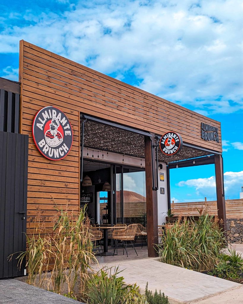 this picture shows Almirante Brunch, a lovely place for lunch in Lajares, Fuerteventura. It has wooden effect cladding on the outside, boho style tables and chairs, plus green plants as you walk in.