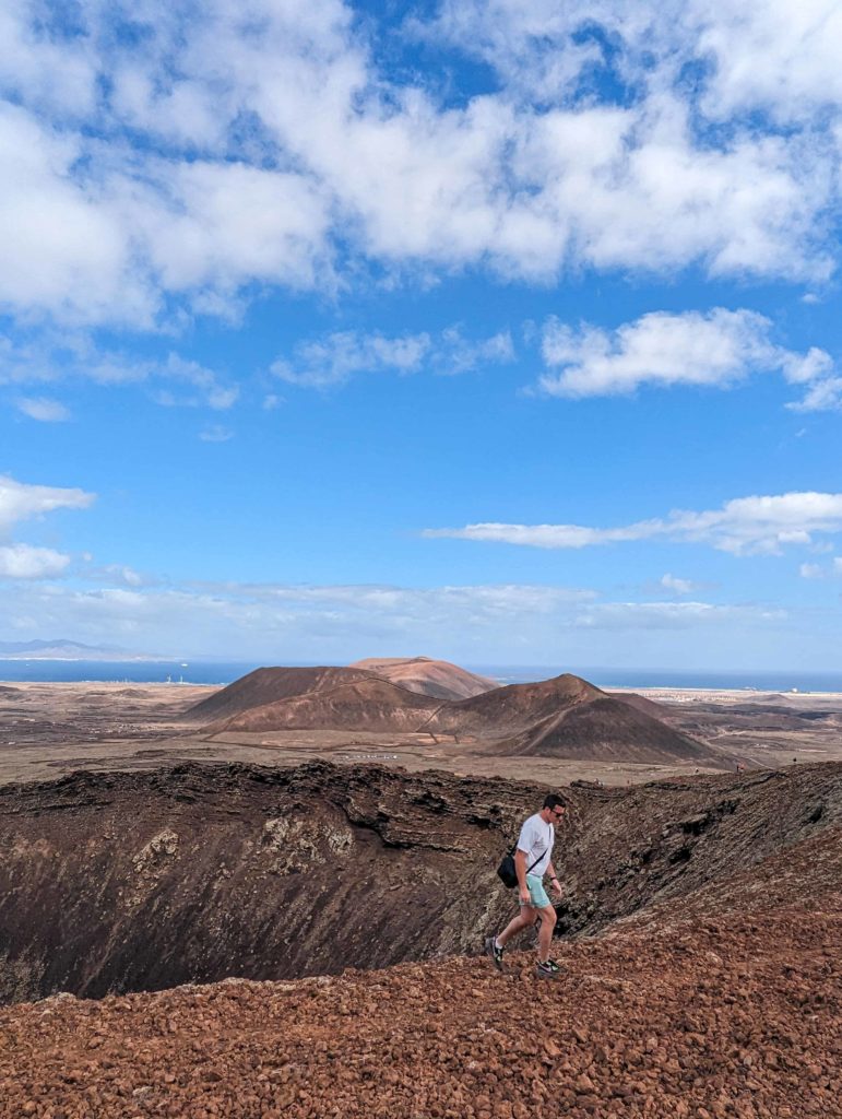 A person stands on the rim of a volcanic crater, photographing the landscape overlooking the ocean and distant mountains under a blue sky in Fuerteventura.