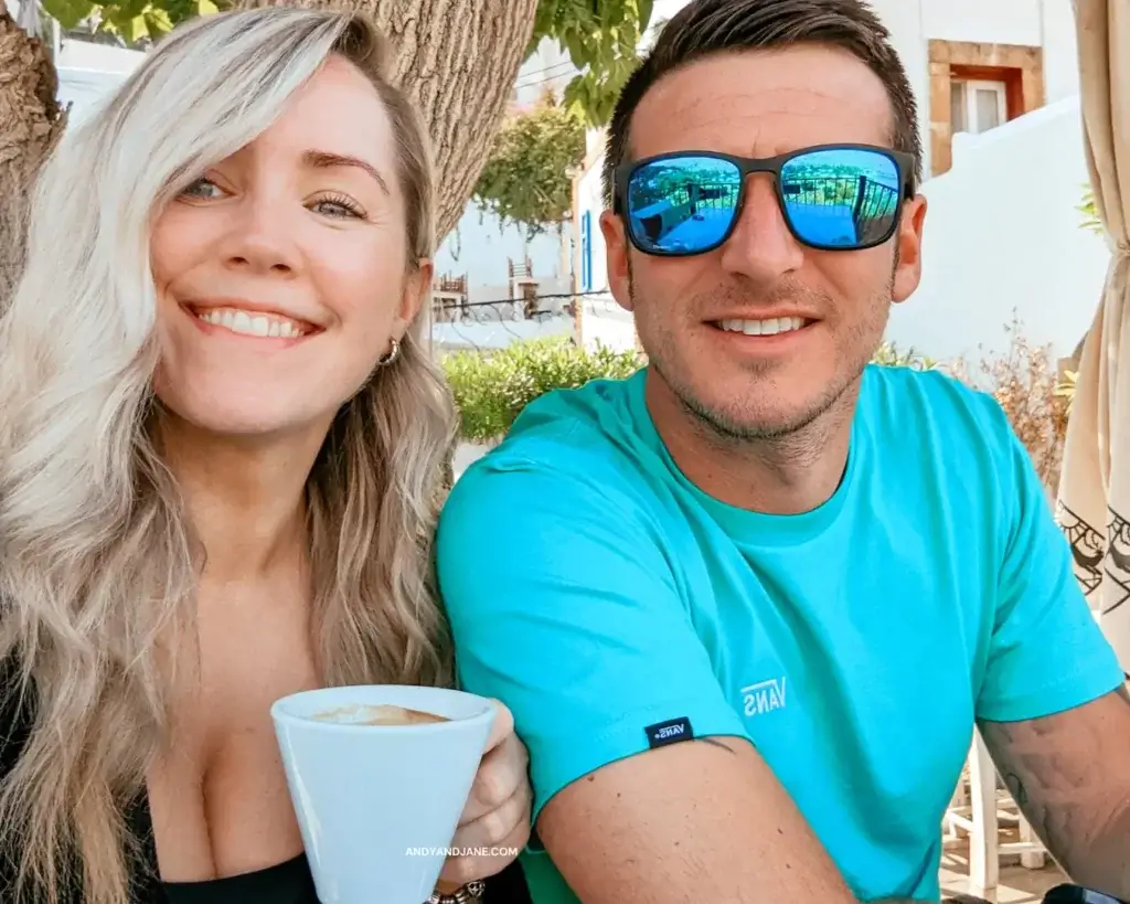 Andrew & Jane smiling into the camera with their coffees in hand at Rainbird Bar in Lindos.