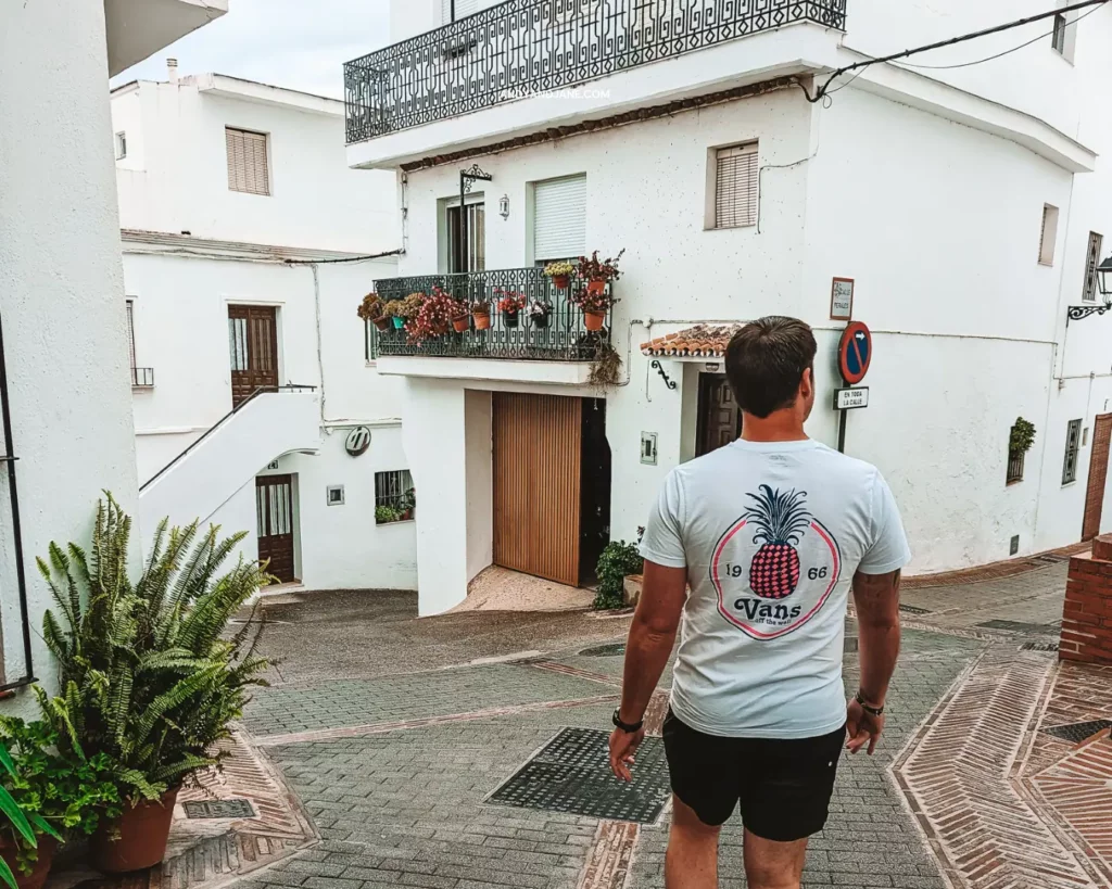 A man walking through a whitewashed village in Spain, passing houses with flower pots handing on the balcony & wearing a pineapple tshirt.