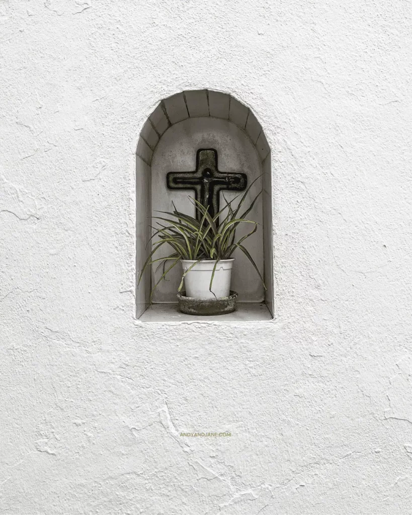 An alcove built into the side of a house with a small flower pot & cross behind it.