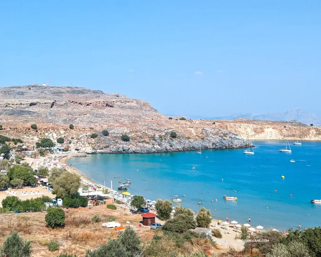 Lindos Beach - a golden sandy beach with crystal clear waters overlooking a bay & out to the sea with small boats dotted all across the water.