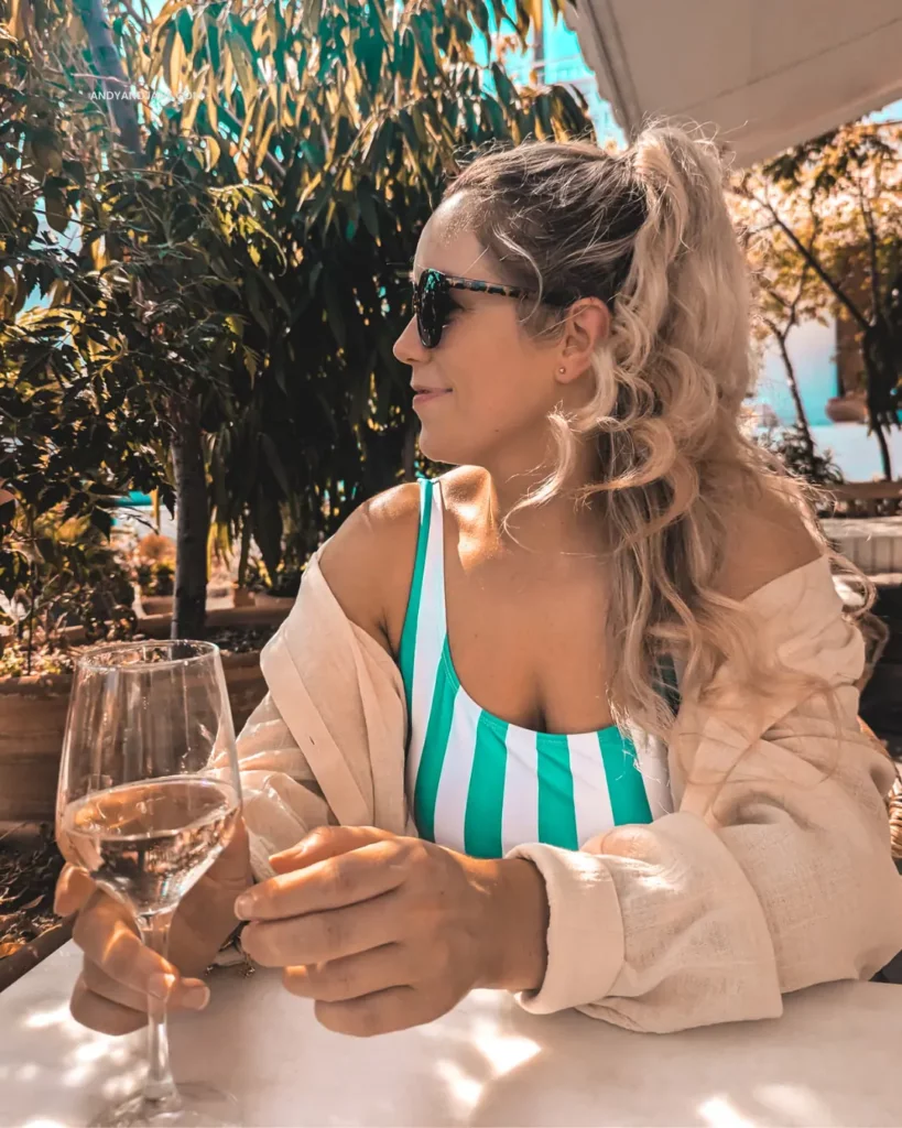 Jane wearing a cream oversized shirt & stripy green swimsuit, looking away from the camera with her glass of white wine in hand