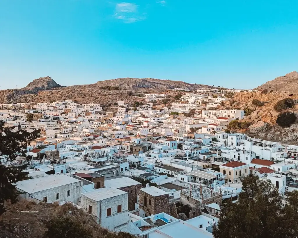 Overlooking the whitewashed buidings & burnt orange rooftops of Lindos Village from the steps of the Acropolis. Mountains & blue skies in the background.