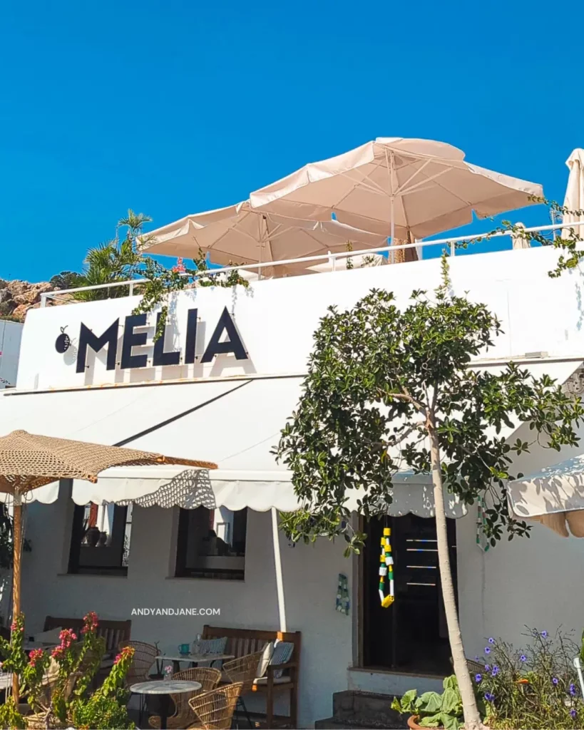 The outside of Melia Cafe in Lindos, a white building with a Melia sign above the outdoor seating area, with umbrellas on the roof top & a blue sky above.