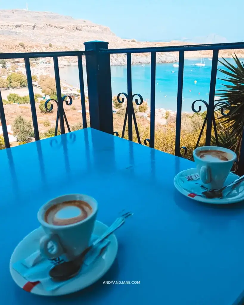 Two coffees sitting on a blue table overlooking the beach & the bay in Lindos.