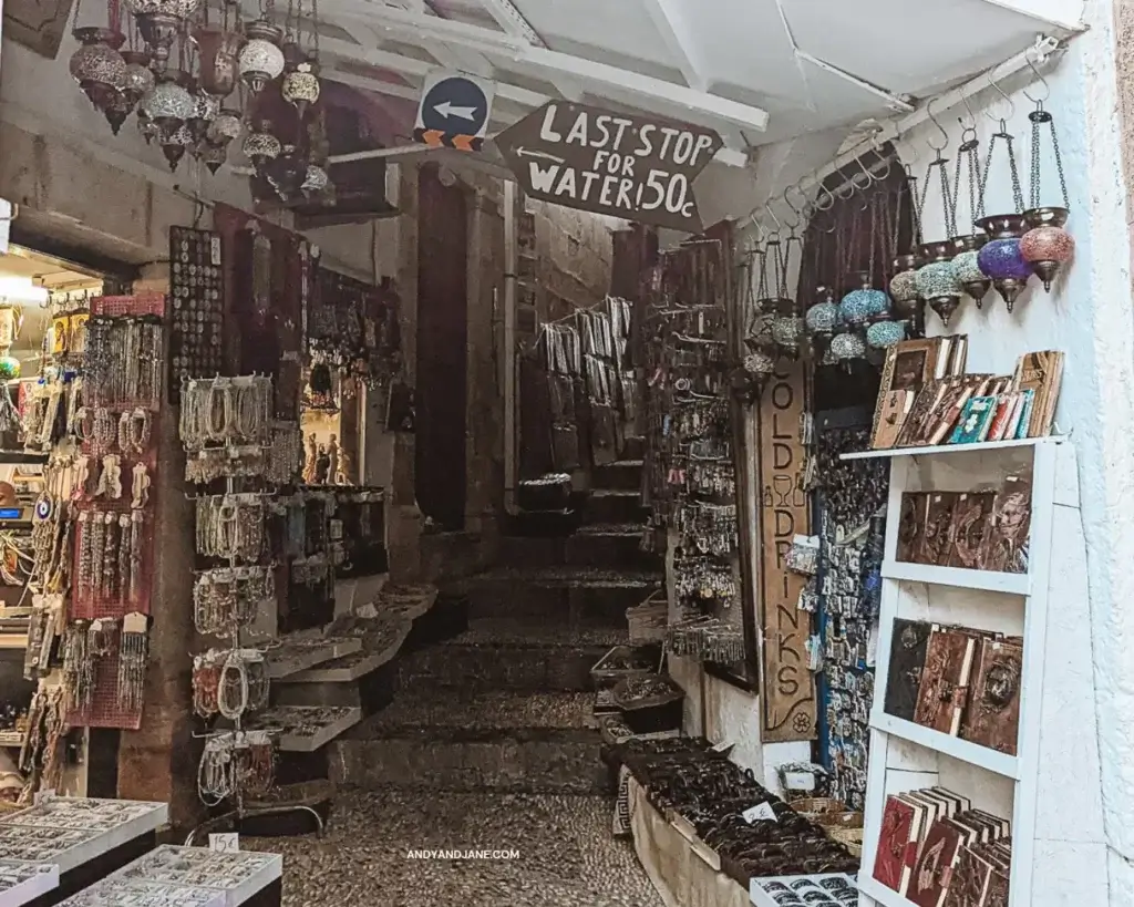 A small alleyway in Lindos village with souveniers, arts & crafts lining the walls. A sign above says 'Last Stop for Water' - as the steps lead to the Acropolis on Lindos.