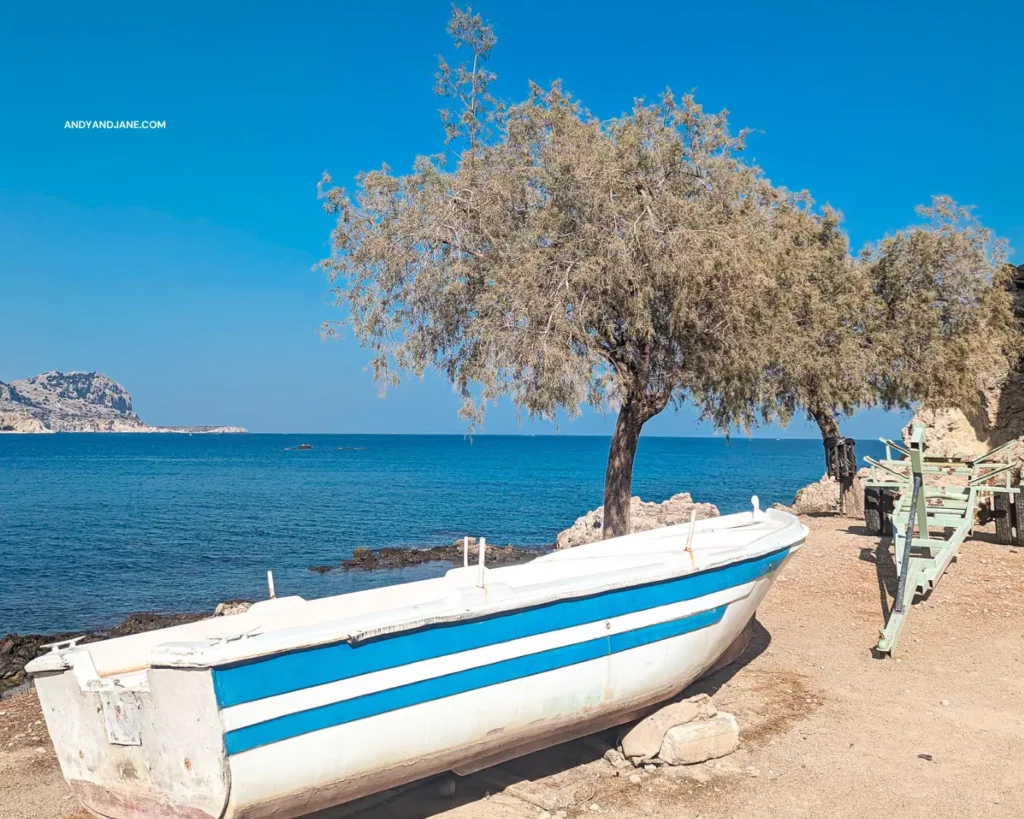 A boat with blue & white stripes, parked next to a tree & the ocean in Stegna Harbour.