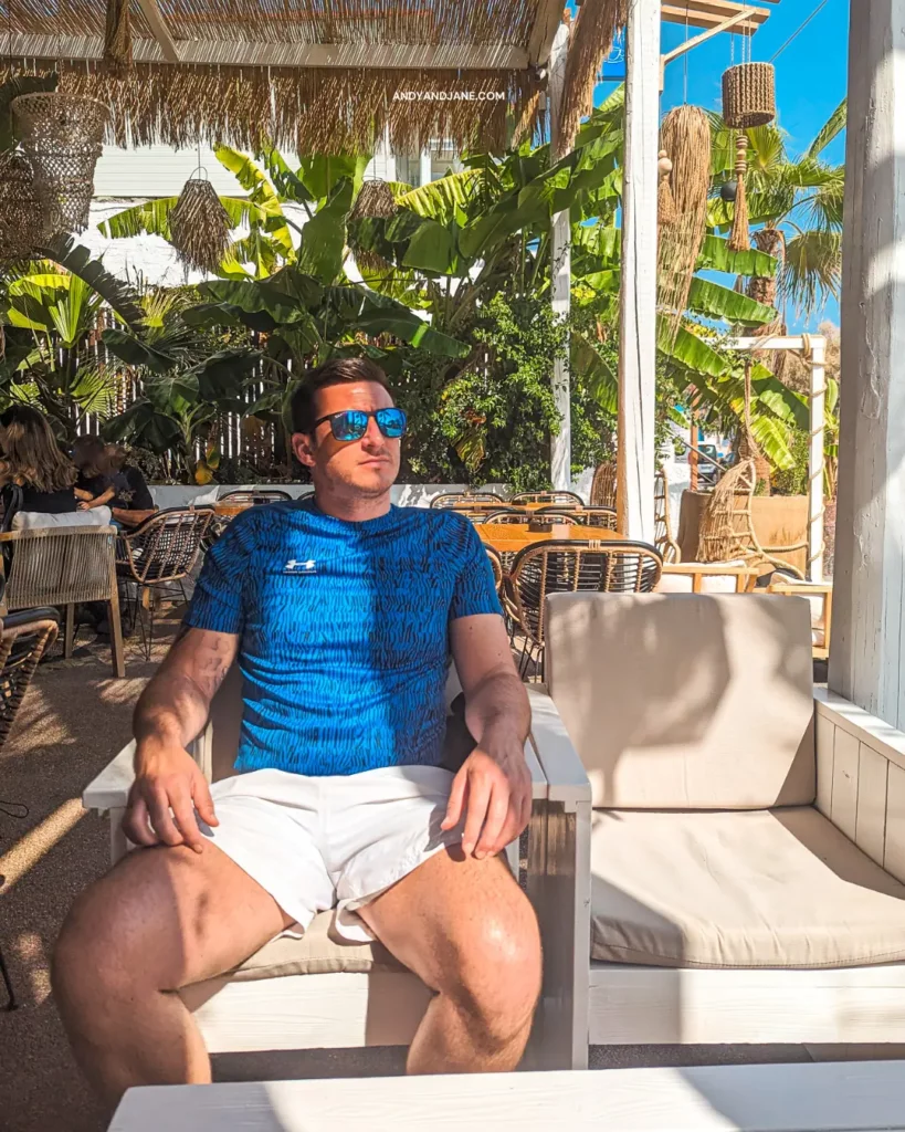 A man sitting on a comfy chair at a beach bar in a blue t shirt & sunglasses.