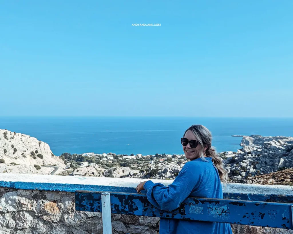 Jane sitting on the blue bench at Stegna Viewpoint in Rhodes, wearing a blue shirt & dark sunglasses smiling back to the camera.