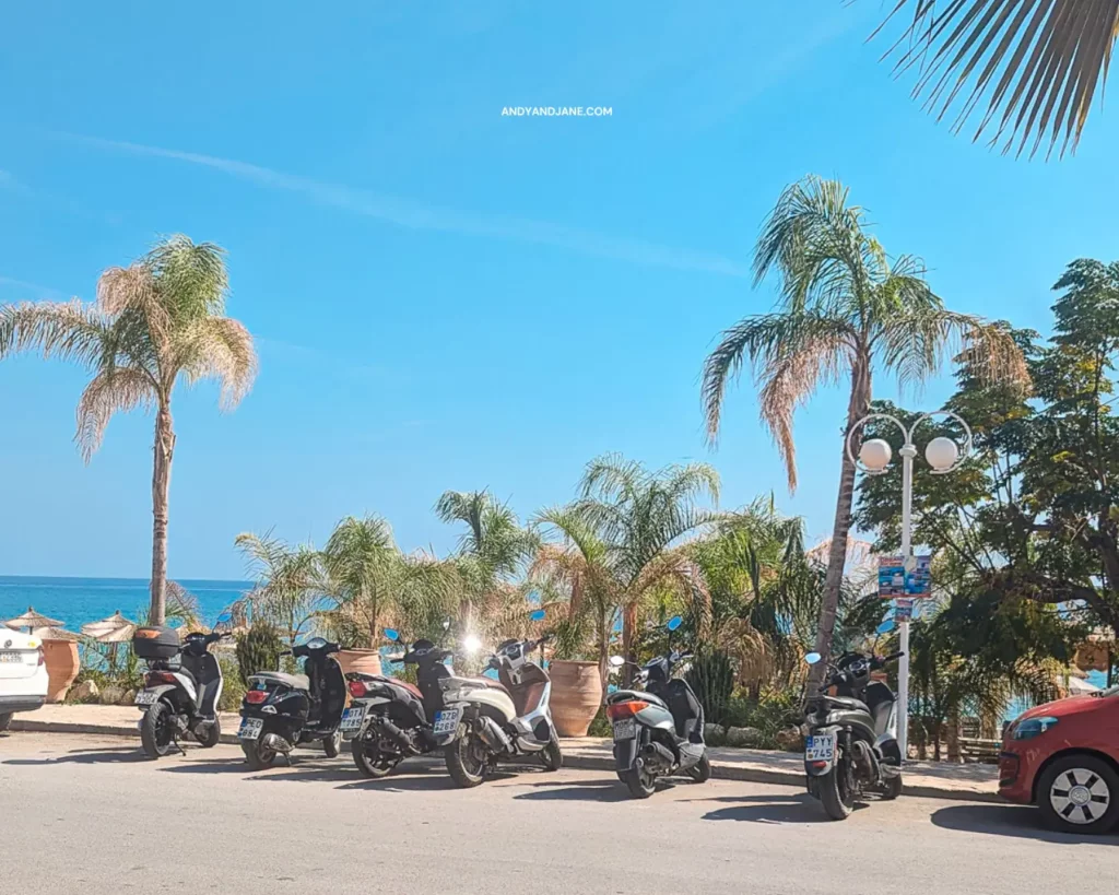 Motorcycles neatly parked by the ocean at Stegna Beach, offering a picturesque view of the sea & the palm trees above.