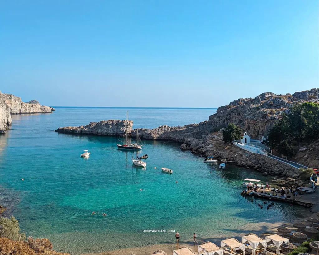 How To Spend One Day In Lindos - St.Pauls Bay - a beautiful blue bay with cliffs & rock formations surrounding it. A white chapel sits to the right with a path leading up to it. Boats float in the middle of the water.