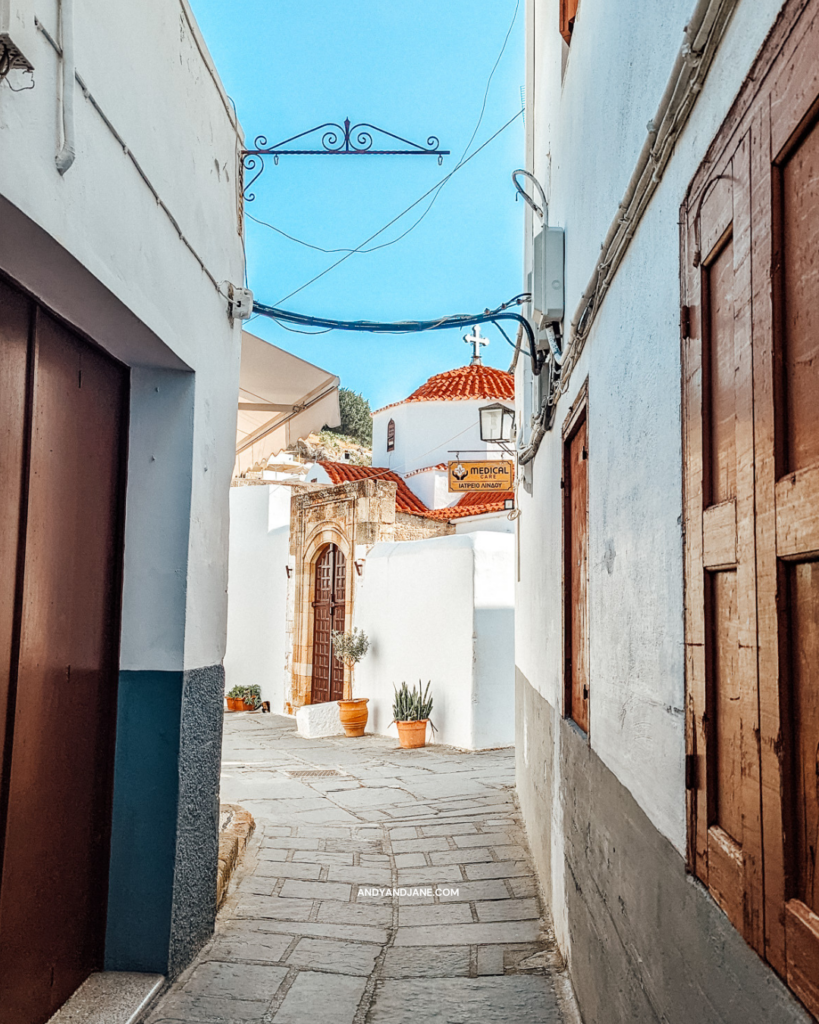 A view down one of the streets in Lindos towards a church with a cross on top & burnt orange coloured roof.