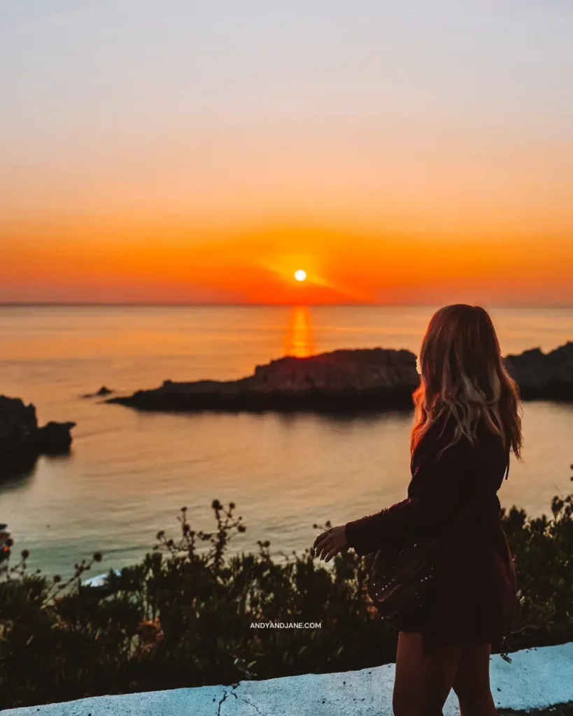 Jane standing on top of St.Paul's Bay in front of the sun rising creating an orange & yellow sky.