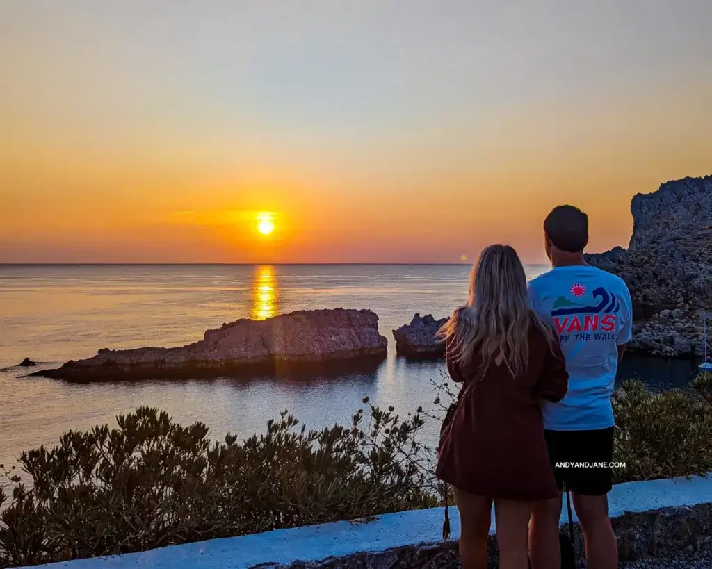 Jane and Andrew watching the sunrise at St.Pauls Bay in Lindos. They are standing beside each other linking arms looking at the orange skies.