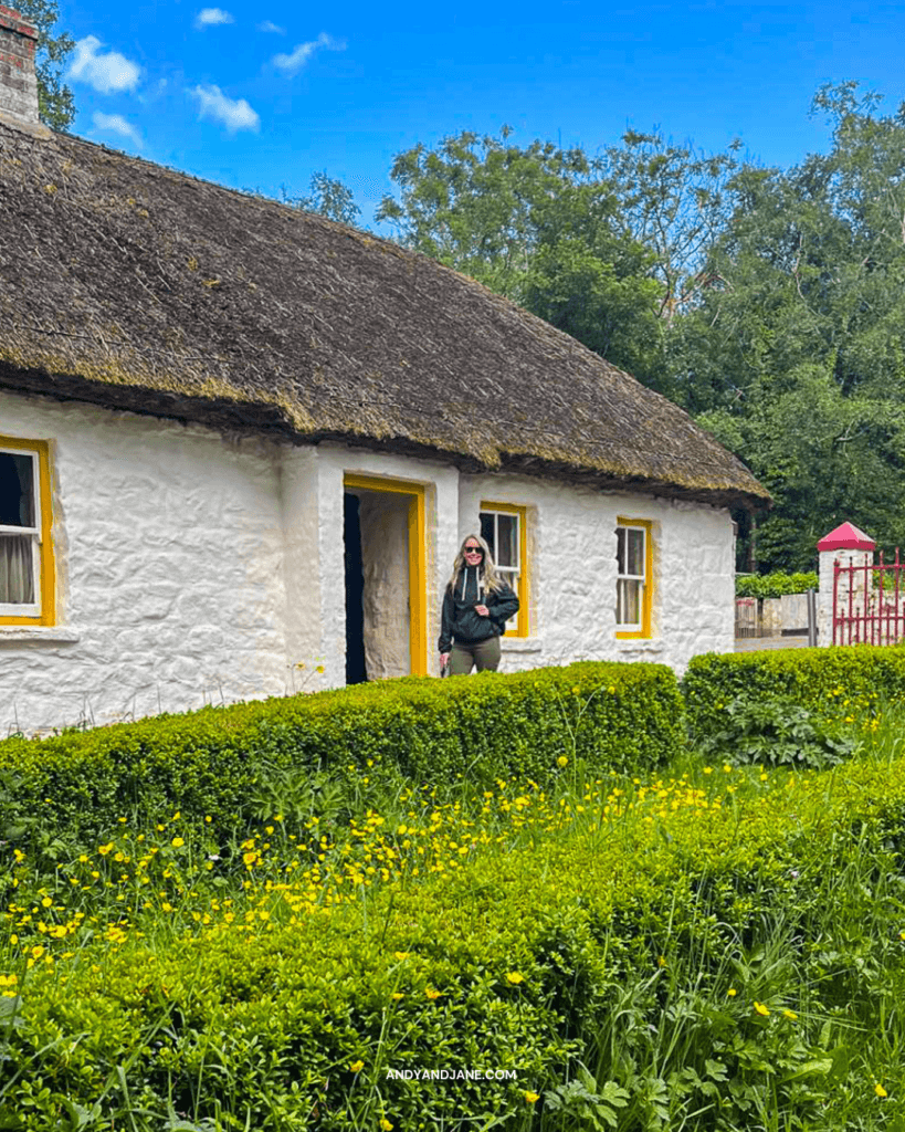 Jane standing at the door of a thatched cottage with yellow window frames & a green garden out front.