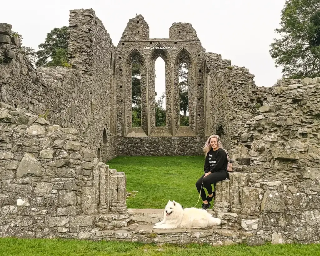 Jane sitting on a wall at Inch Abbey in front of the ruins - with Luka the white Samoyed dog at her feet.