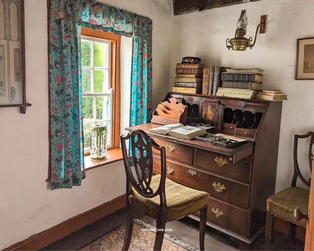 A room inside one of the old houses at the museum, with a dark mahogany desk & chair, filled with books & sitting beside a small window with blue flowered curtains. 