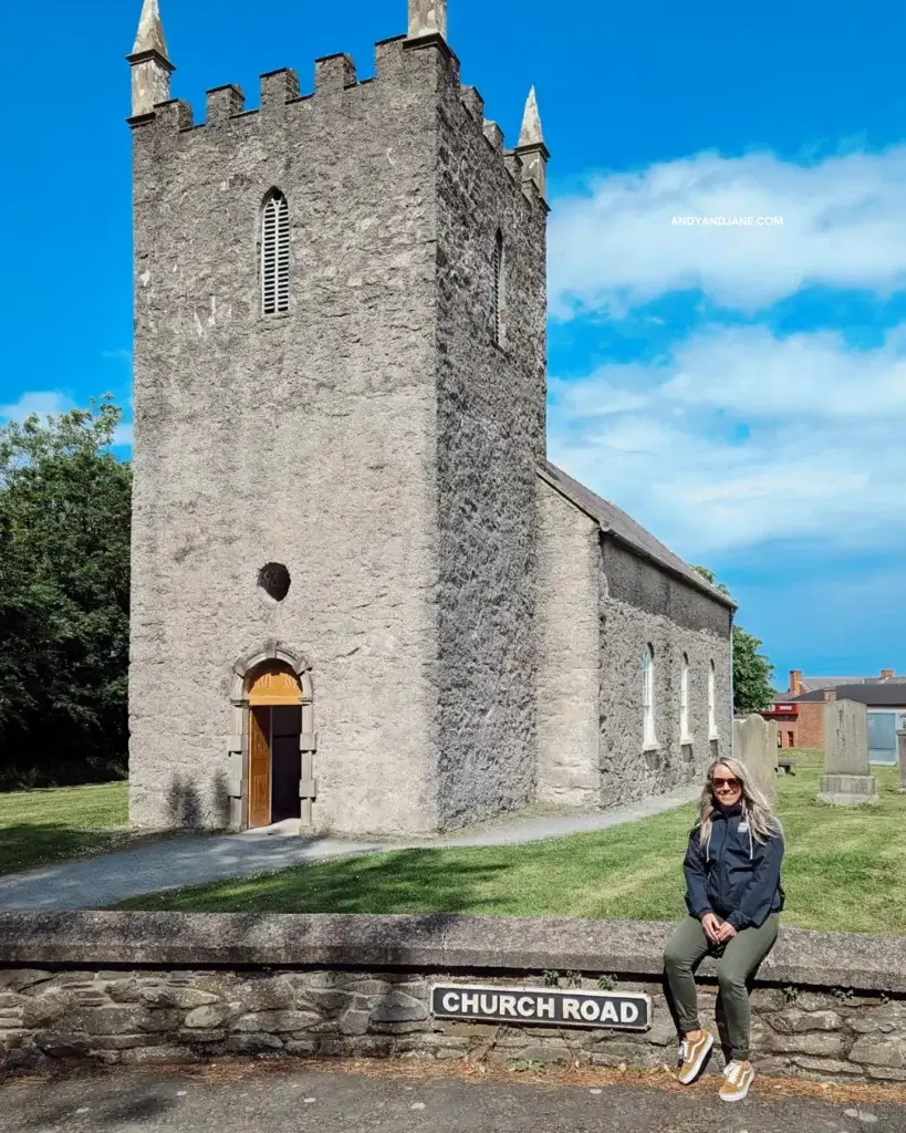 Jane sitting on a low wall in front of the Kilmore Church of Ireland. The sign on the wall says Church Road & there are headstones in the graveyard behind. 