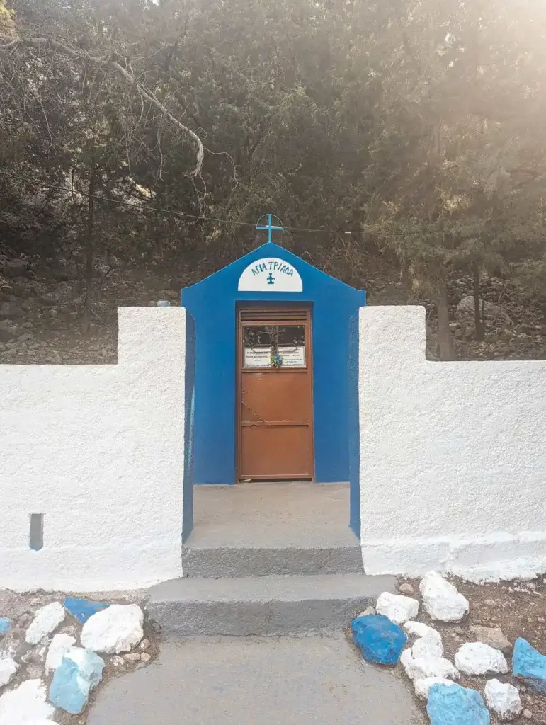 Small blue and white chapel entrance with a cross above the door, surrounded by trees and painted stones on the ground.