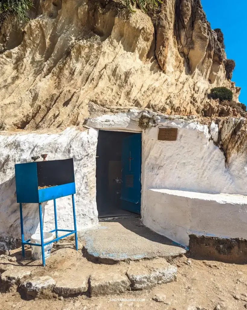 A small chapel built into the side of a rock with a blue door and white seat outside.