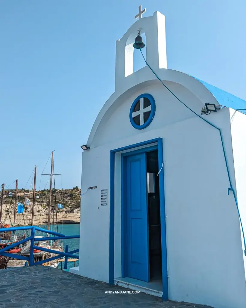 A tiny white chapel with a blue door & small white cross above it. Sitting in a harbour with boats beside it.