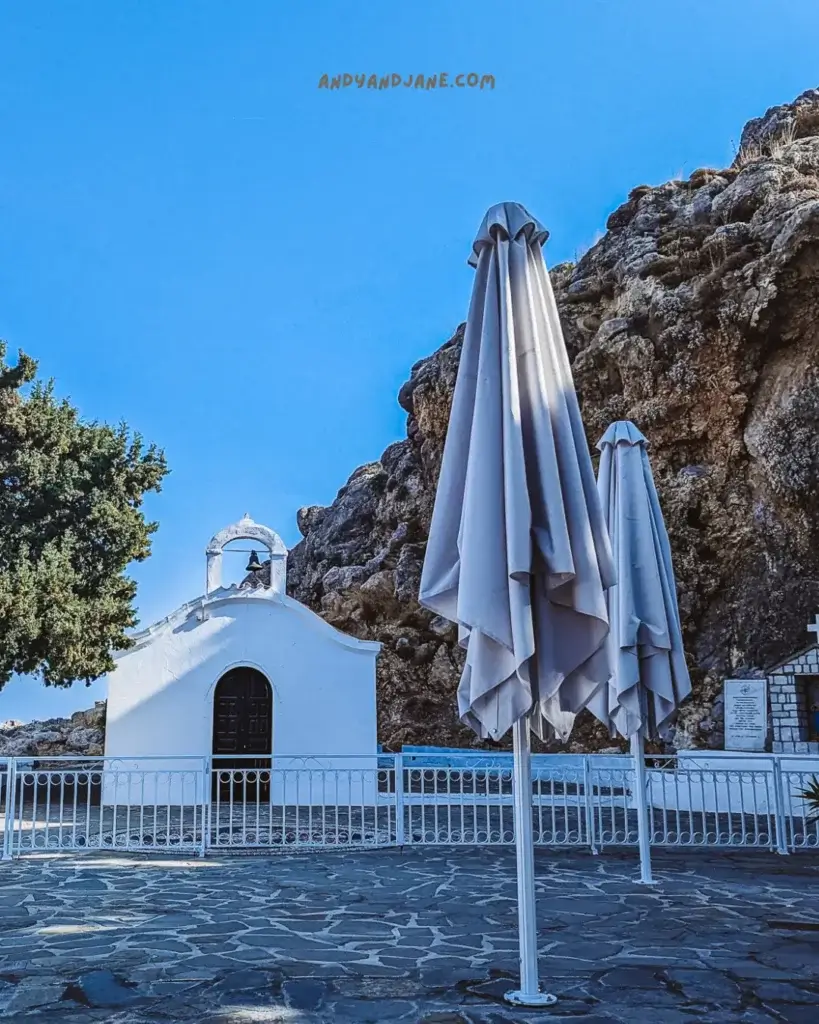 A small white church underneath some rocks with a white gate surrounding it & two large umbrellas in front.