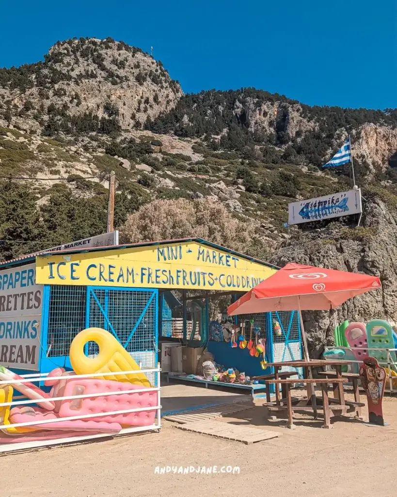 A mini-market at Tsambika Beach with a yellow sign saying - Ice Cream, Fresh Fruits & Cold Drinks. Inflatables  for the sea sit out the front of the shop beside a small seating area.