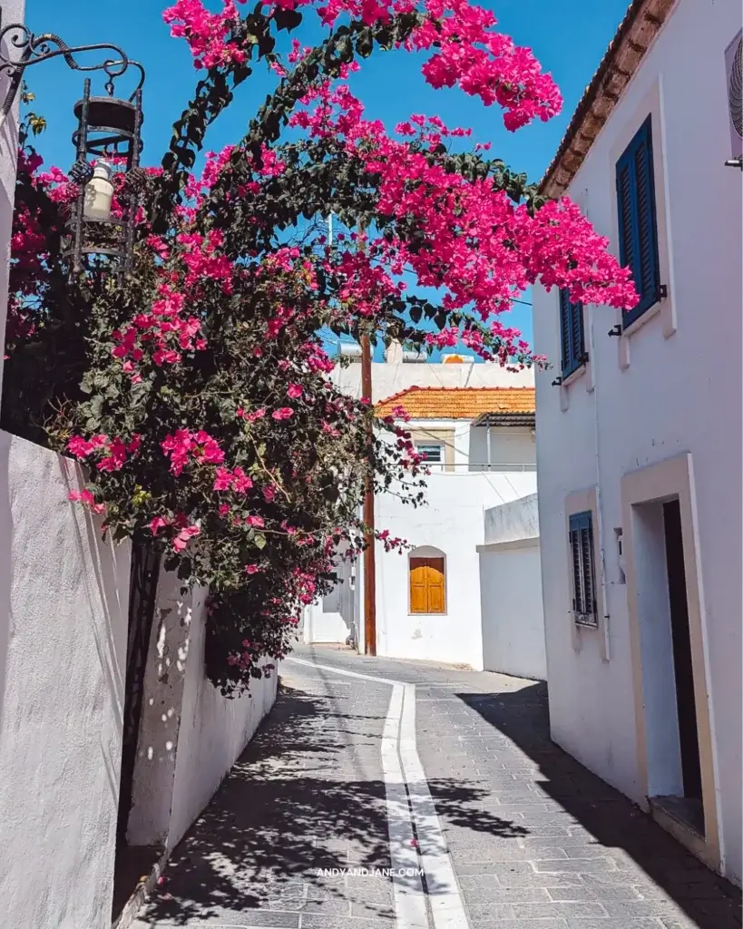 A quiet street in Gennadi, white building with pine shutters & an orange roof. Pink flowers reach over the street.