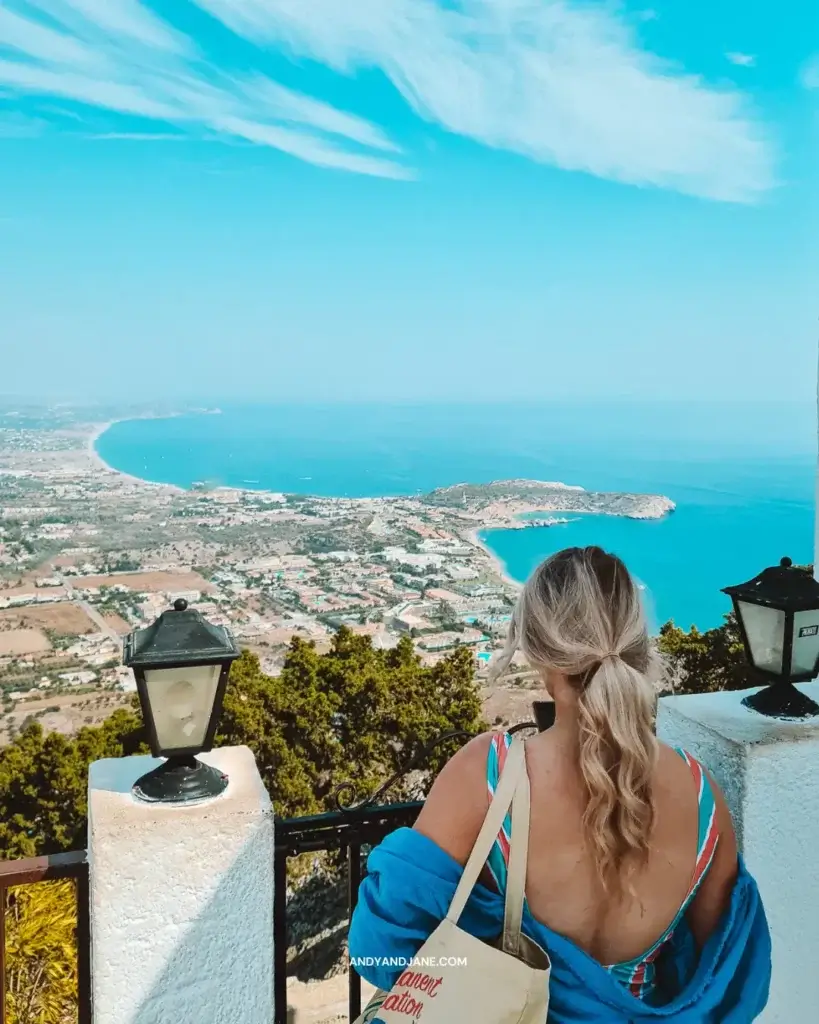 A girl looking out over a white wall towards views of the sea & nearby village of Kolymbia.