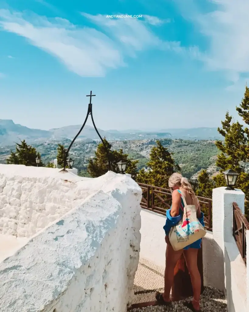 A girl walking around the white wall of the Monastery with a cross on top with views across the mountains of Rhodes.