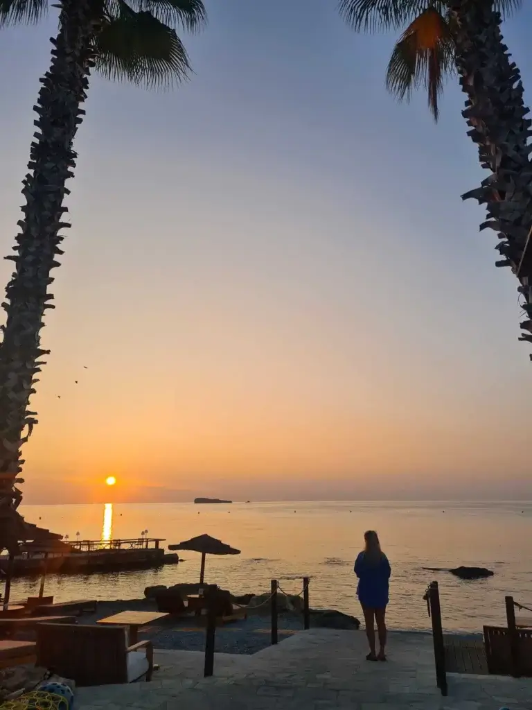 A girl standing on a beach looking out to the sun rising in the sky.