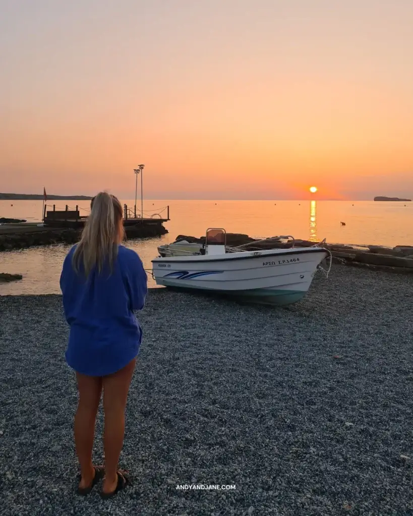 A girl standing in front of a small fishing boat watching the sun come up on the beach.