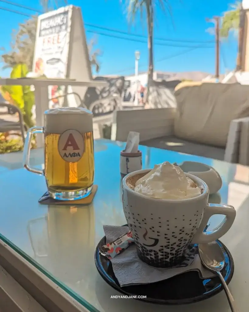 A coffee topped with cream & a cold beer on a table looking out to the blue sky in the street.