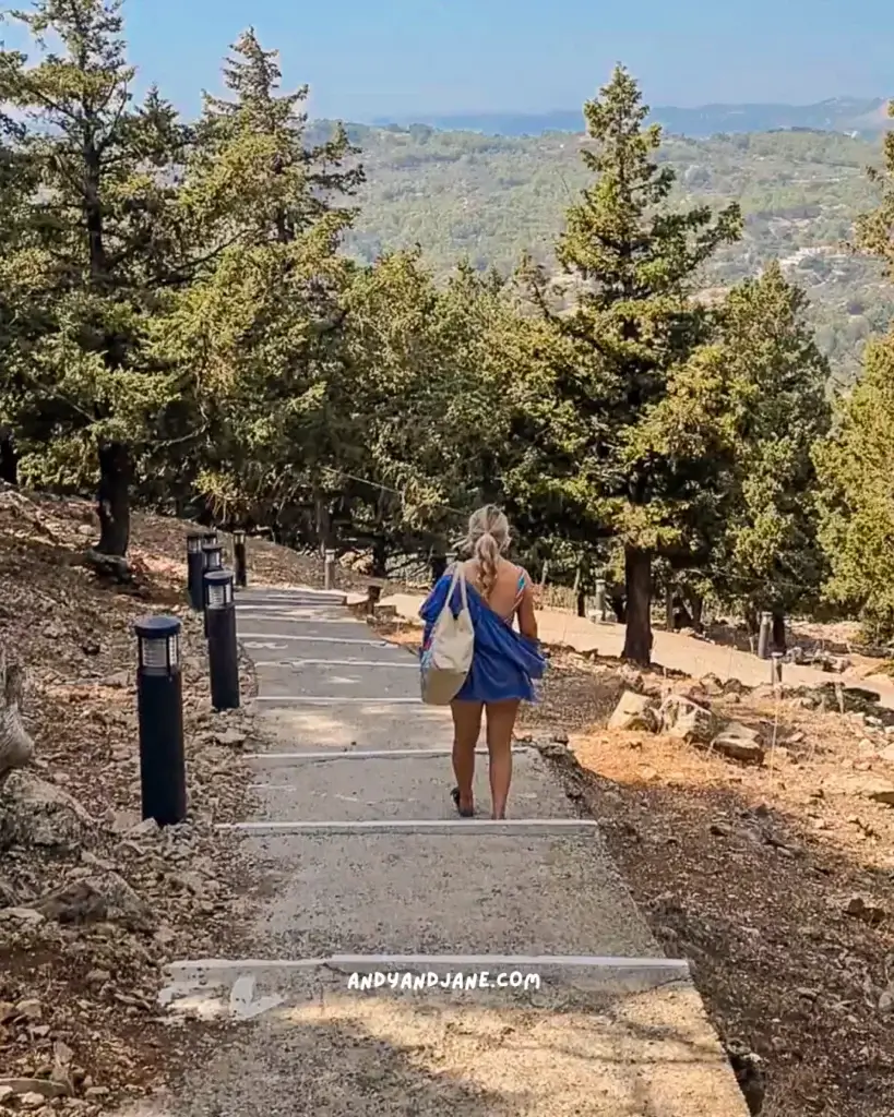 A girl wearing a blue shirt & carrying a beach bag descending the steps from Tsambika Monastery through the trees overlooking a mountainous landscape.