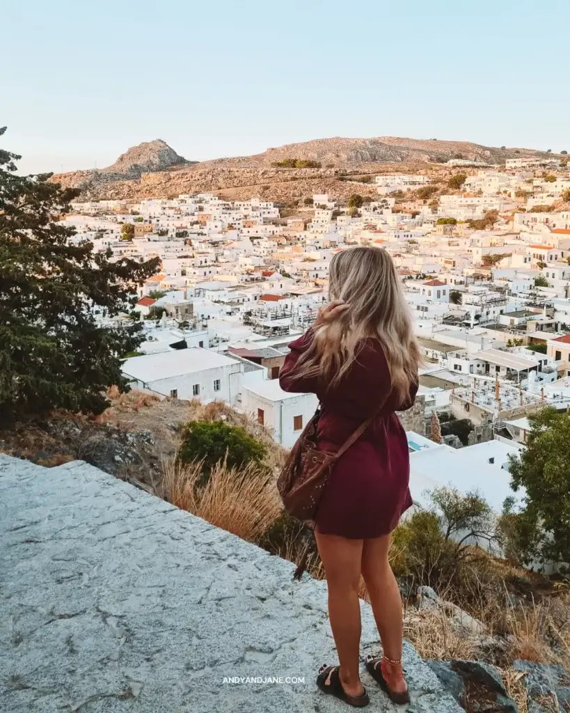 A girl standing on steps in a brown dress, looking over the orange rooftops of the whitewashed village of Lindos.