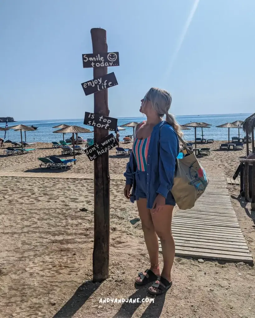 A woman in beach attire stands beside a wooden post with signs reading "Smile today," "Enjoy life," "Is too short," and "Don't worry, be happy" at Tsambika Beach Rhodes, with loungers and umbrellas lining the shore.