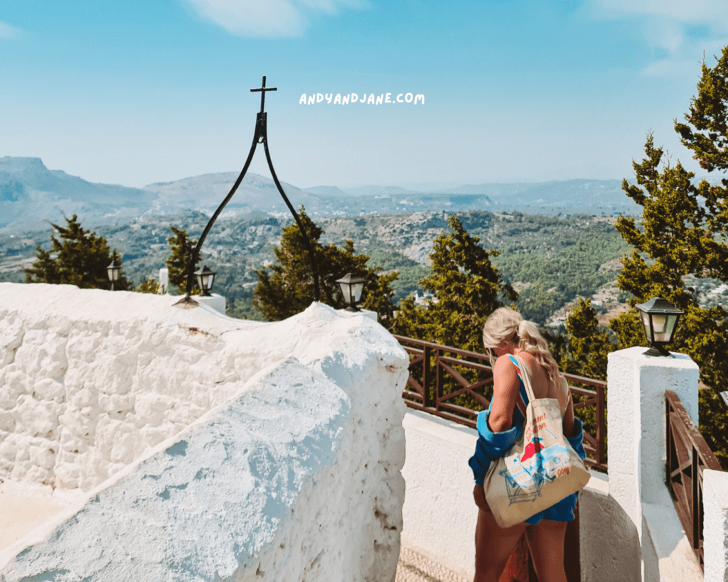 A girl with a beach bag walks along a path near the white stone structure of Tsambika Monastery, which overlooks a mountainous landscape under a clear sky.