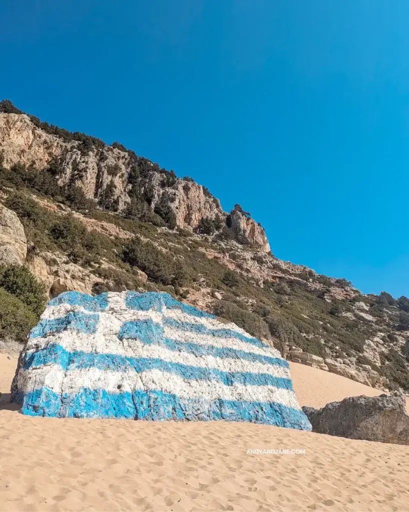 A greek flag painted on a large rock on the beach in front of a cliff.