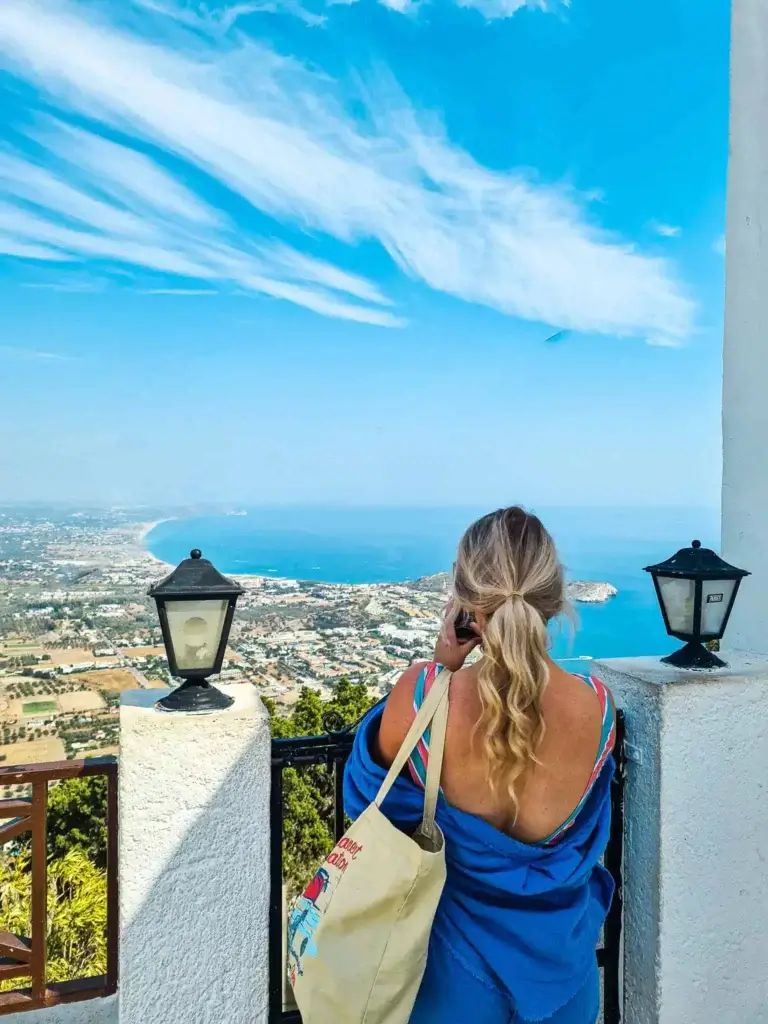 A person with long hair in a ponytail, wearing a blue top and carrying a tote bag, looks out at a coastal landscape across to Kolymbia & Afantou, from a high vantage point between two lanterns.