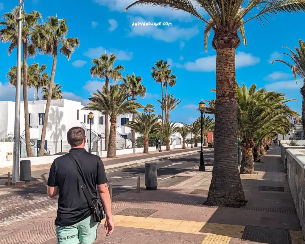 A man in a black shirt & green shorts walking down a street lined with palm-trees in Corralejo.