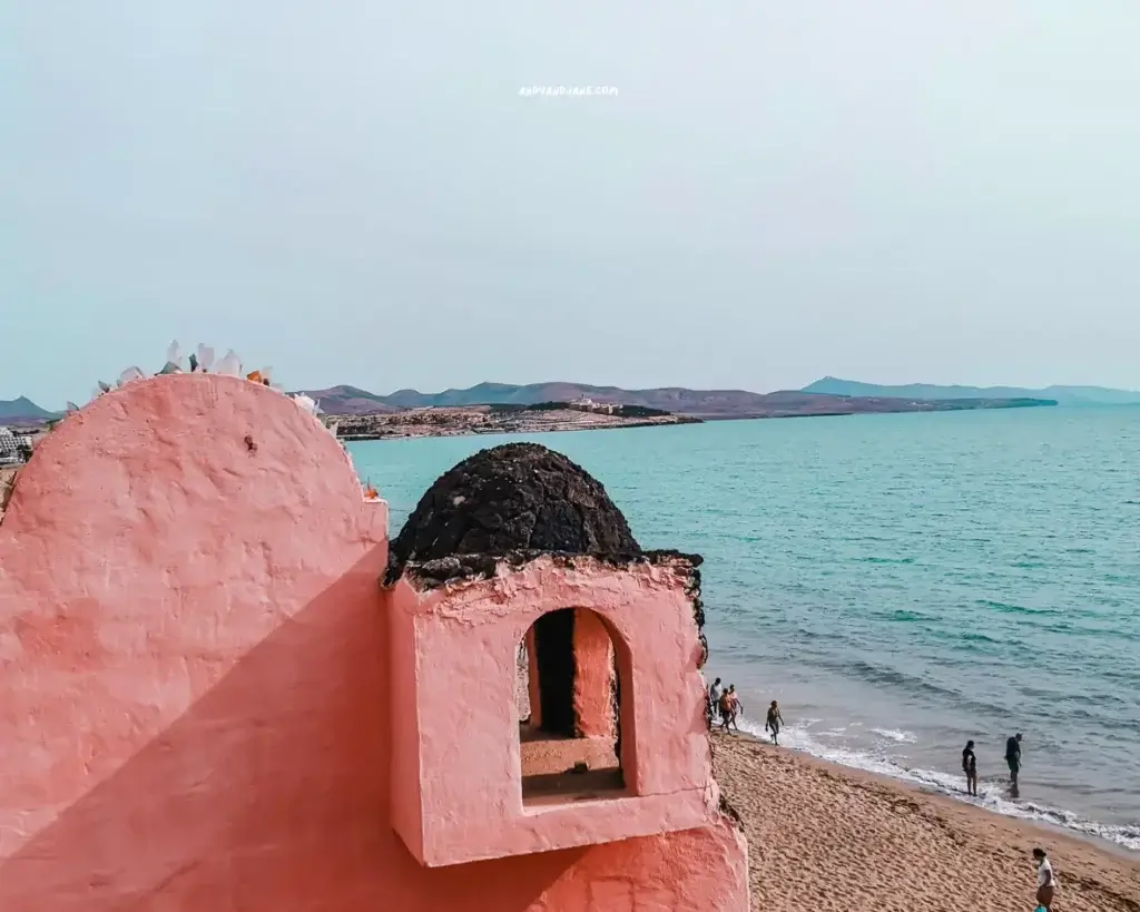 A picturesque pink building on the beach, surrounded by individuals walking along the shoreline, basking in the sun.