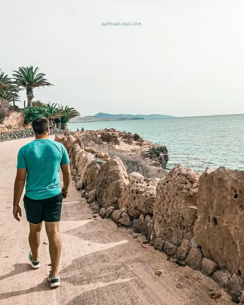 A man leisurely walks on a promenade, with a dramatic rocky wall serving as a backdrop to the serene coastal scene.