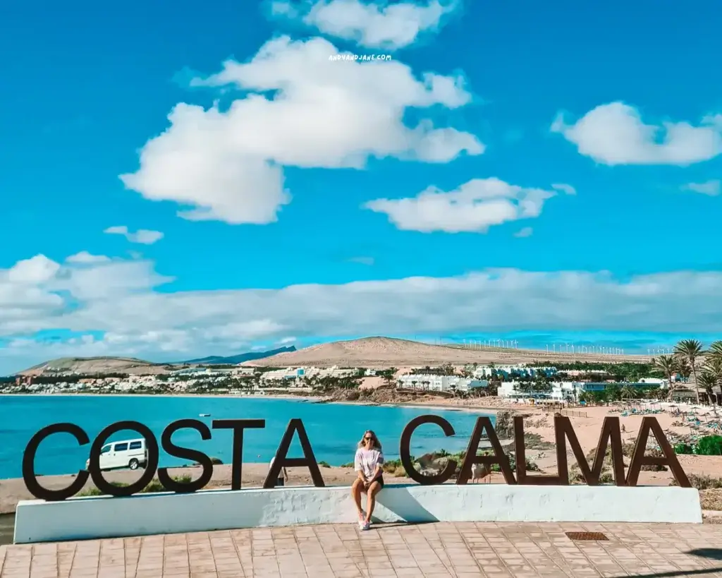 A woman sitting in front of a large metal sign that says 'Costa Calma' with the resort, mountains and beach in the distance behind her.