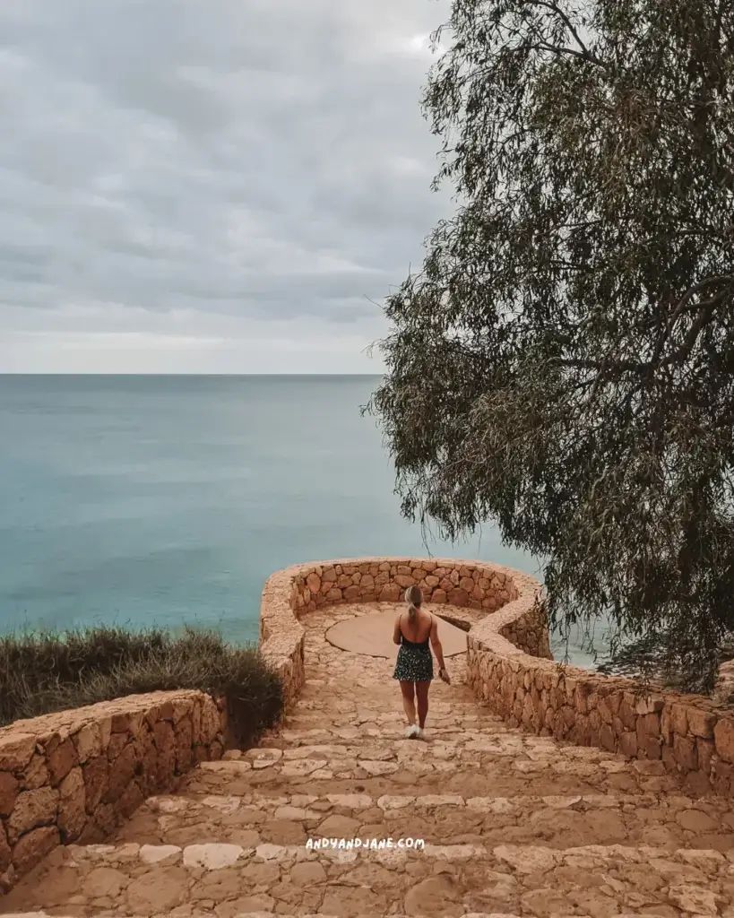 A woman walking down stone steps towards the ocean viewpoint.