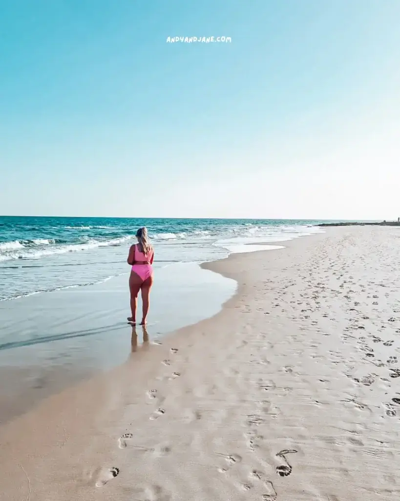 A woman n a pink bikini walking on a white sandy beach, surrounded by the blue ocean.