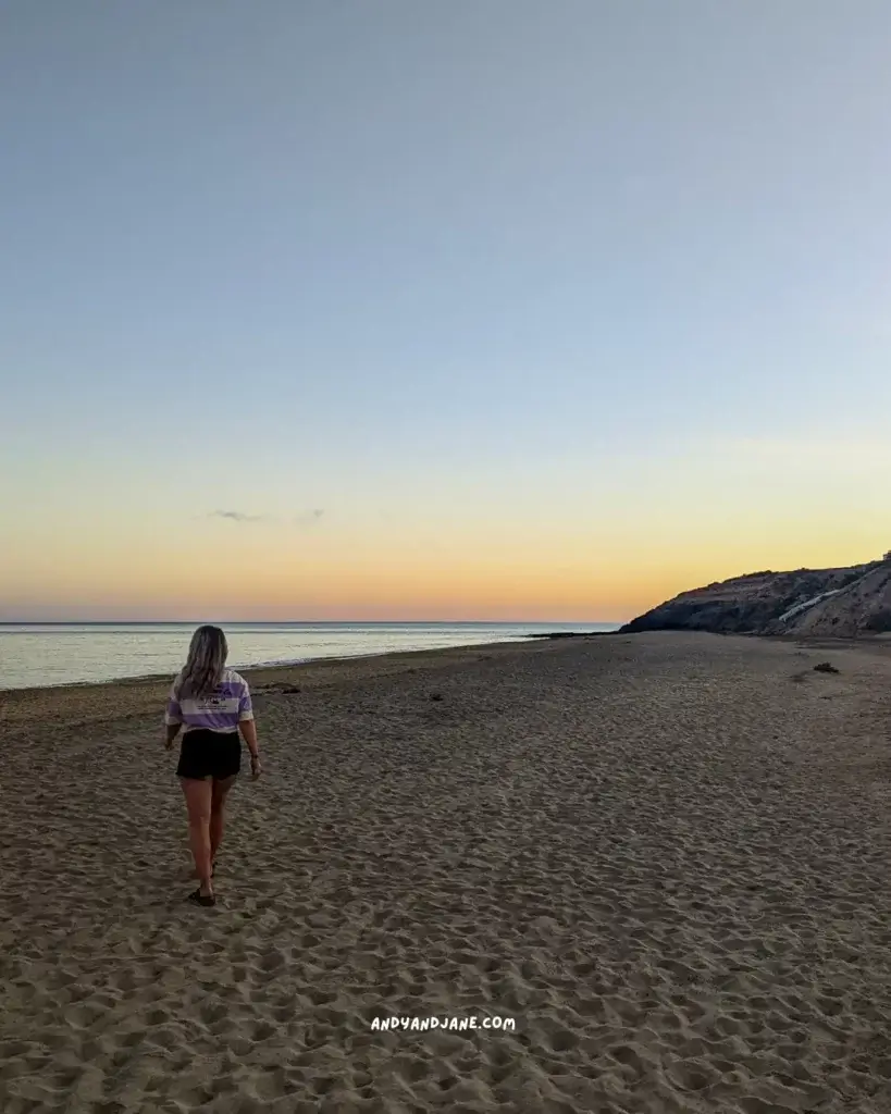 A woman walking along a sandy beach at sunset.