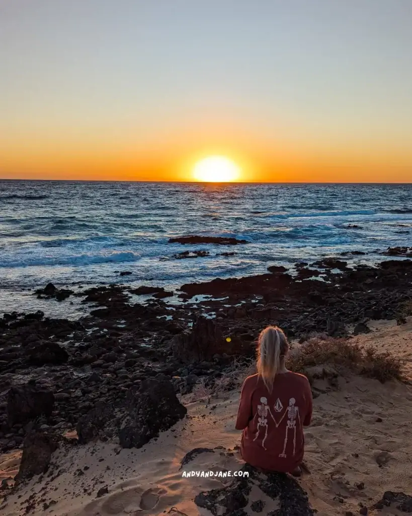 A woman sitting on a rocky beach watching the sunrise on the horizon in Corralejo.